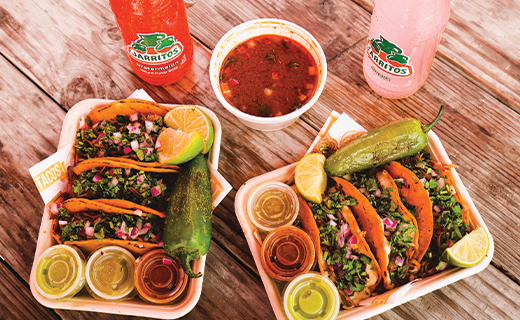 Kiké's Red Tacos Jarritos event showcasing a meal for two. The image features mouthwatering Queso Tacos accompanied by consome, salsas, lime, and peppers.