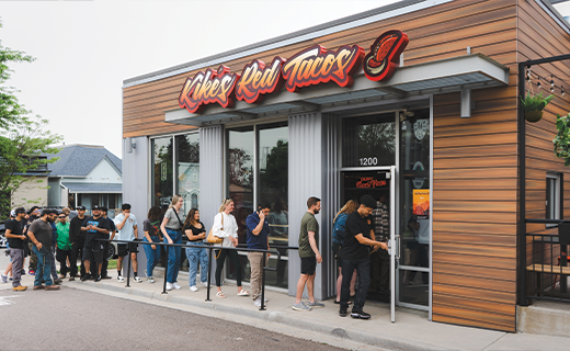 The day of the grand opening at Kiké's Red Tacos brick & mortar, captured in an image showing a long line of enthusiastic and excited customers eagerly awaiting their turn to experience delicious Birria tacos.